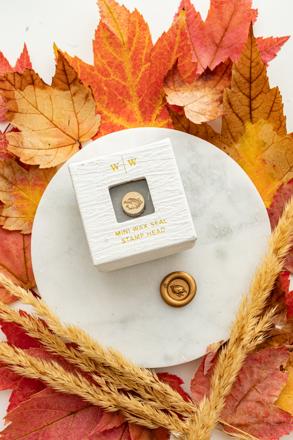 Mini turkey wax seal stamp head in white packaging with gold foil lettering, paired with a gold wax seal impression on a marble surface surrounded by autumn leaves and wheat stems.