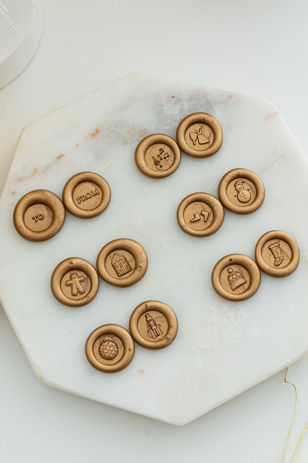 Flat lay of gold wax seals showcasing all twelve Holiday Mini Wax Seal Stamp designs, including festive icons like a snowman, nutcracker, stocking, and “TO” and “FROM” imprints, displayed on a marble slab.