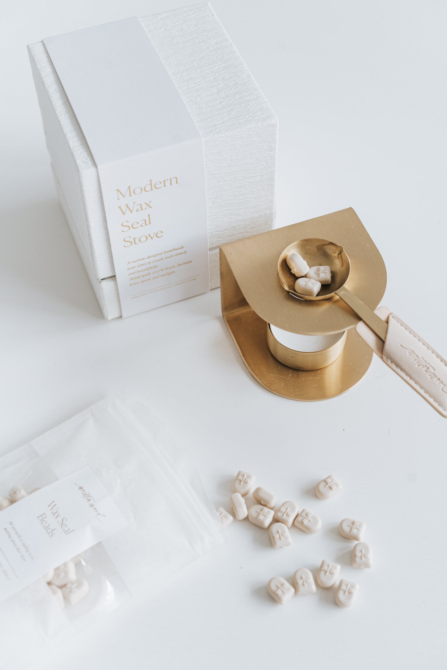 Brass wax seal stove with a melting spoon holding ivory wax beads, placed beside its textured white packaging and a scattered pouch of wax beads
