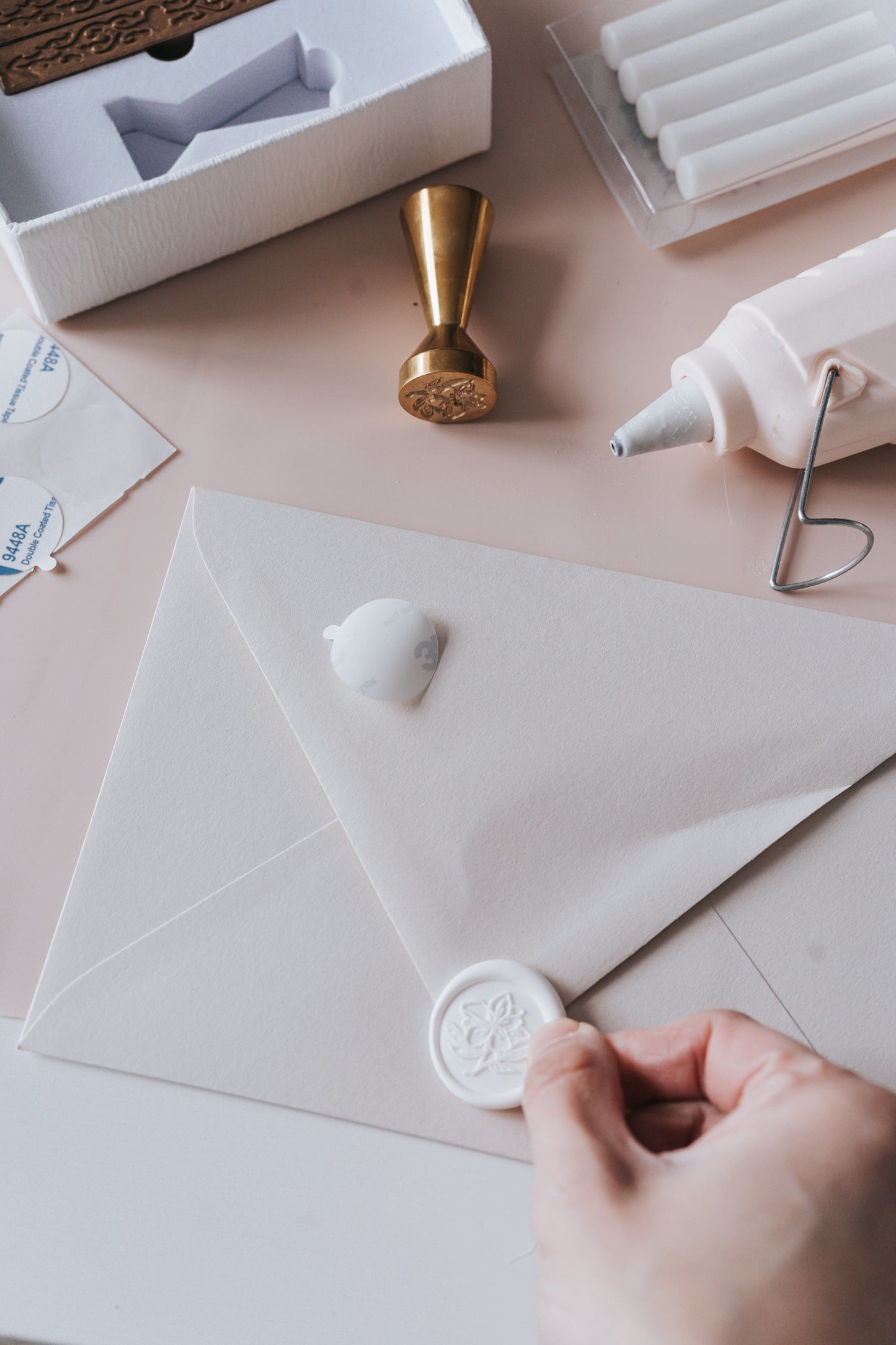 A hand applying a white wax seal with a floral design to an envelope, with wax seal supplies, glue gun, and brass stamp nearby.