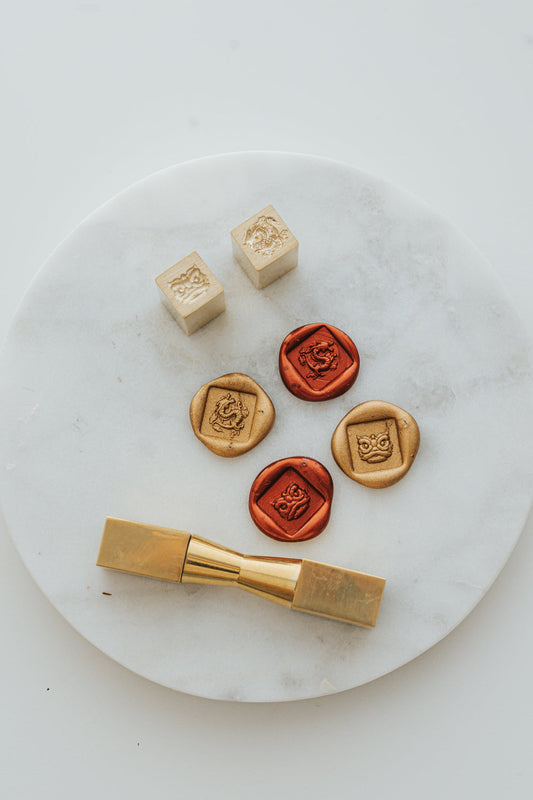 Square Mini Wax Seal Heads with Lion Dance and Dragon Designs with a gold and red wax seal on a white round marble coaster