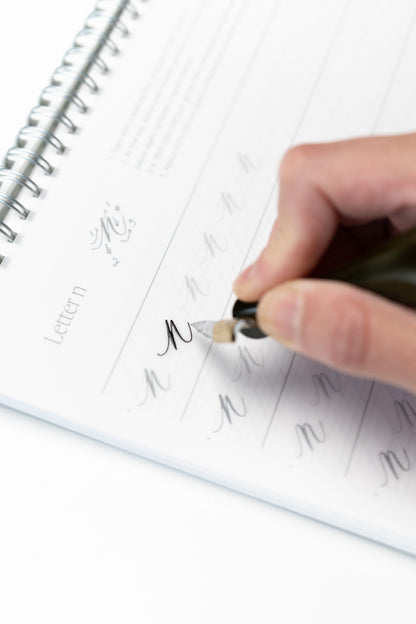 Hand practicing calligraphy with a dip pen on a spiral-bound workbook featuring guided tracing sheets for learning letterforms