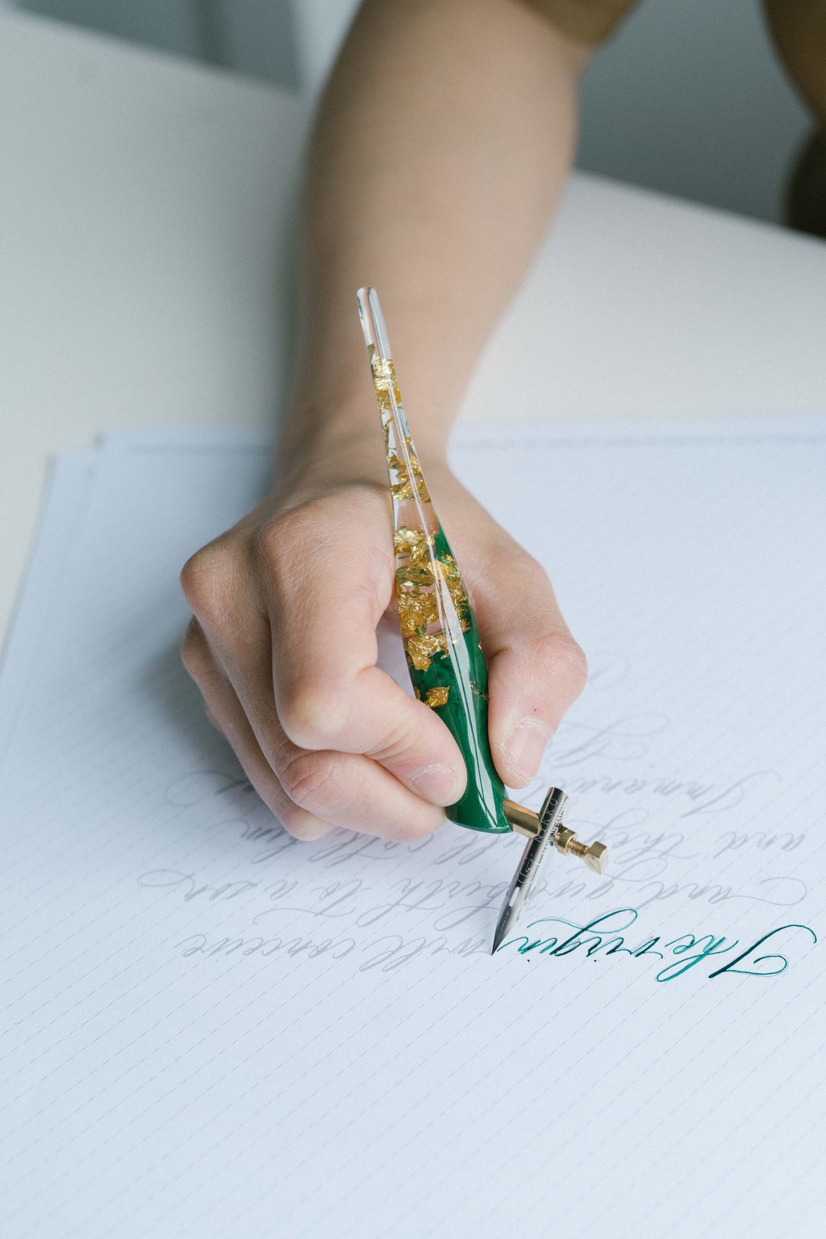 Hand using green oblique calligraphy pen with gold flakes writing modern calligraphy on lined practice paper