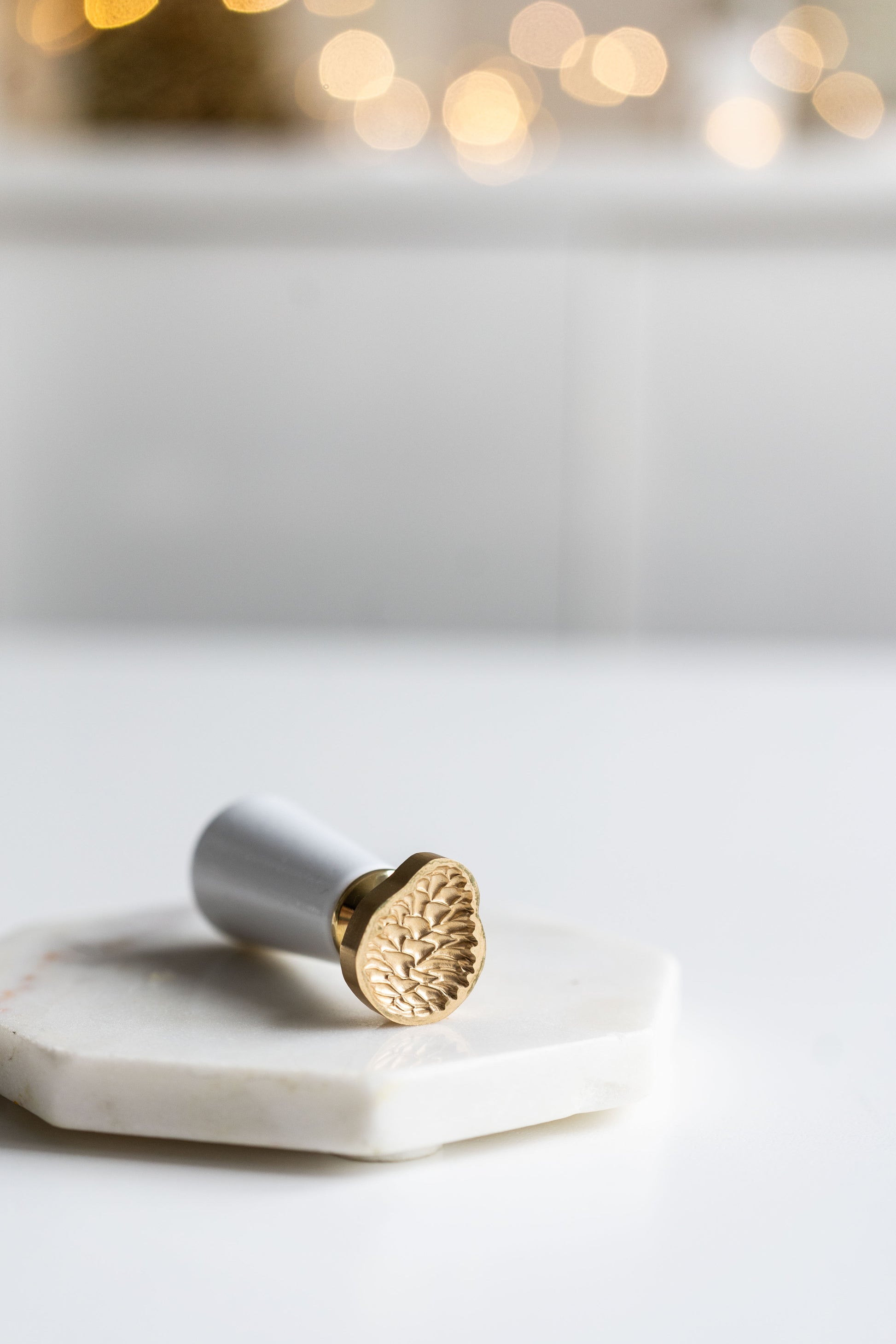 A 3D pinecone wax seal stamp with wooden handle on a marble coaster surrounded by festive lights