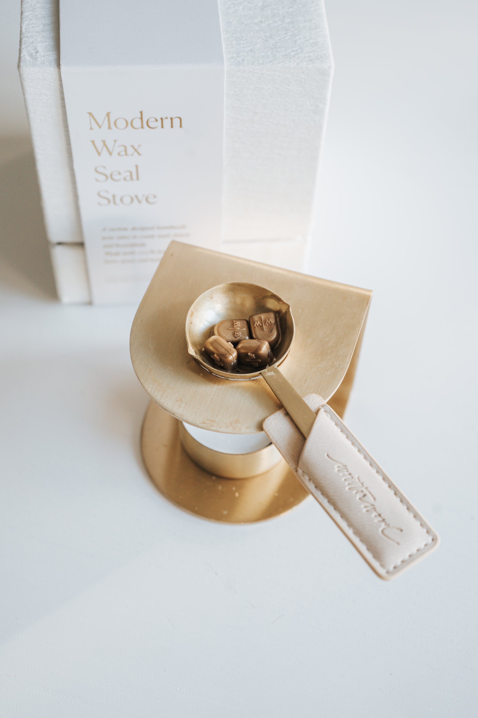 Brass wax seal stove with a melting spoon containing partially melted bronze wax beads, placed beside its elegant white packaging.