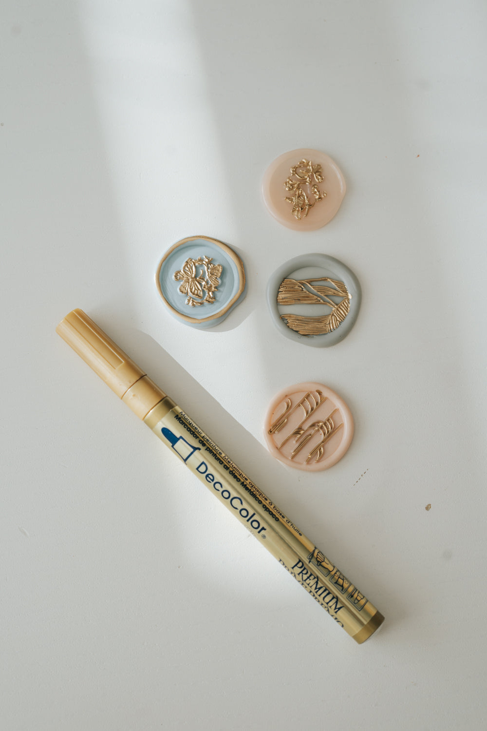 Gold paint pen with four wax seals in pastel tones featuring gold floral and botanical designs on a white surface with soft shadows.