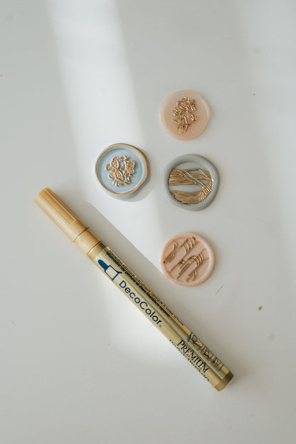 Gold paint pen with four wax seals in pastel tones featuring gold floral and botanical designs on a white surface with soft shadows.