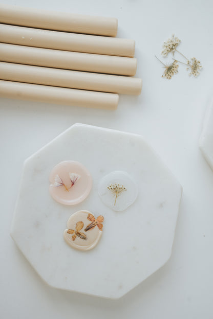 Three handmade wax seals with embedded dried flowers on a white marble tray, next to beige sealing wax sticks and small floral sprigs.