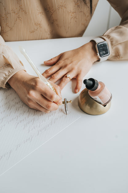 Handwriting elegant calligraphy with a resin pen and blush ink, accompanied by a tilted brass inkwell holding the ink bottle