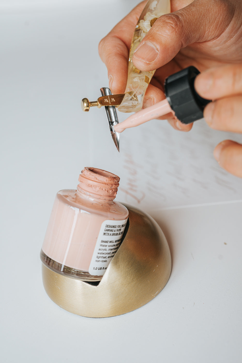 Hand filling a resin pen nib with blush pink ink from a bottle held in a modern tilted inkwell with a sleek faux brass finish