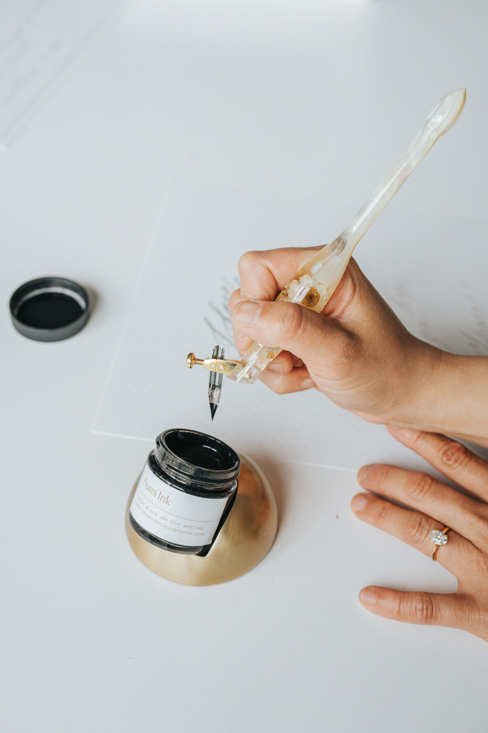 Hand using a resin pen to dip into black sumi ink held by a modern tilted inkwell with a faux brass finish, ideal for calligraphy