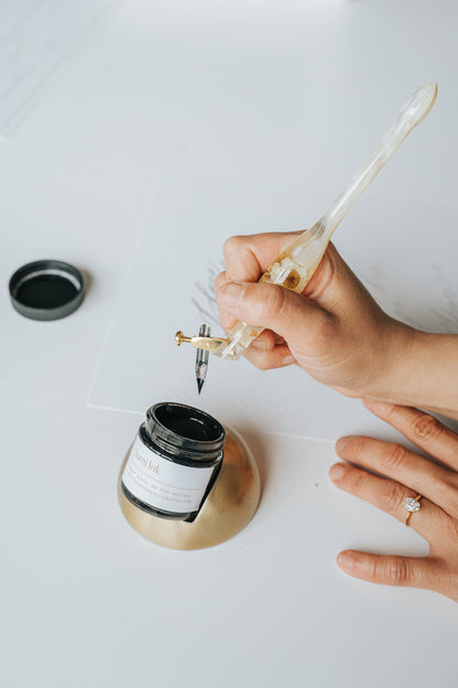Hand using a resin pen to dip into black sumi ink held by a modern tilted inkwell with a faux brass finish, ideal for calligraphy