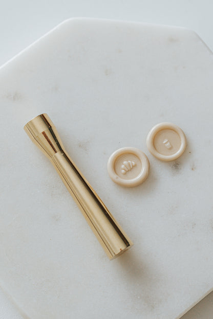 Gold miniature wax seal stamp with croissant and coffee cup designs, placed on a white marble surface