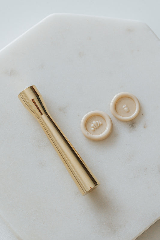 Gold miniature wax seal stamp with croissant and coffee cup designs, placed on a white marble surface