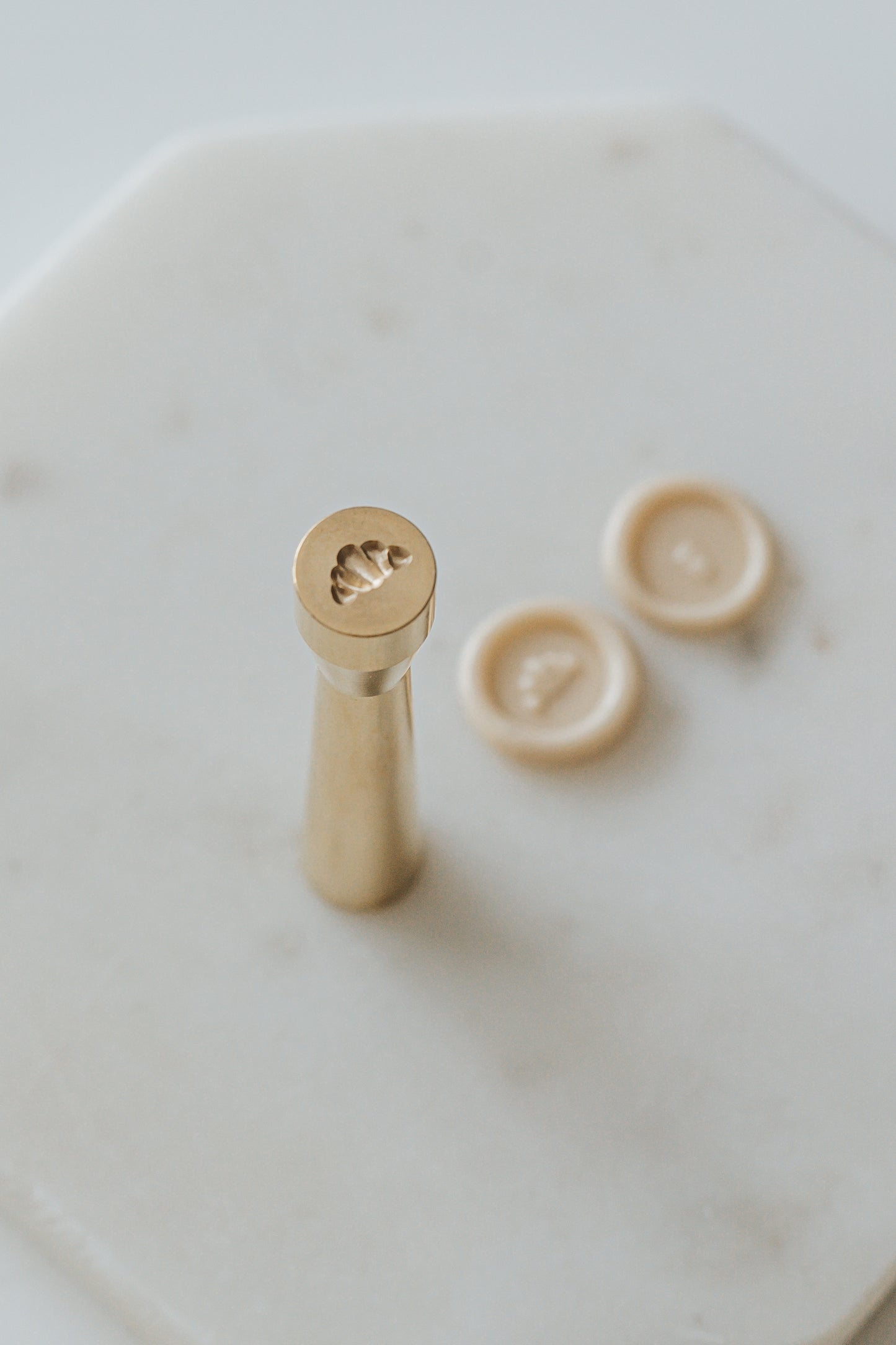 Gold miniature wax seal stamp with croissant designs, placed on a white marble surface with matching beige wax seal