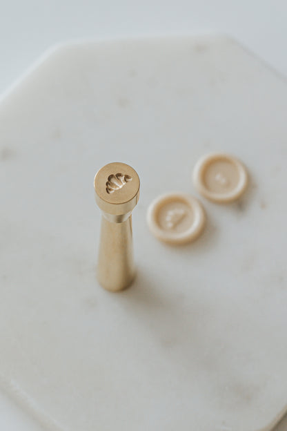 Gold miniature wax seal stamp with croissant designs, placed on a white marble surface with matching beige wax seal