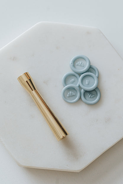 Gold miniature wax seal stamp with croissant and coffee designs, placed on a white marble surface with matching blue wax seal