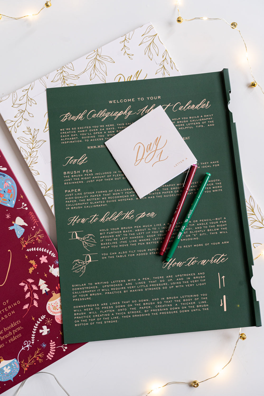 Open Brush Calligraphy Advent Calendar showing a green instruction panel with gold foil text, “Day 1” booklet, and two brush pens in red and green surrounded by warm fairy lights.