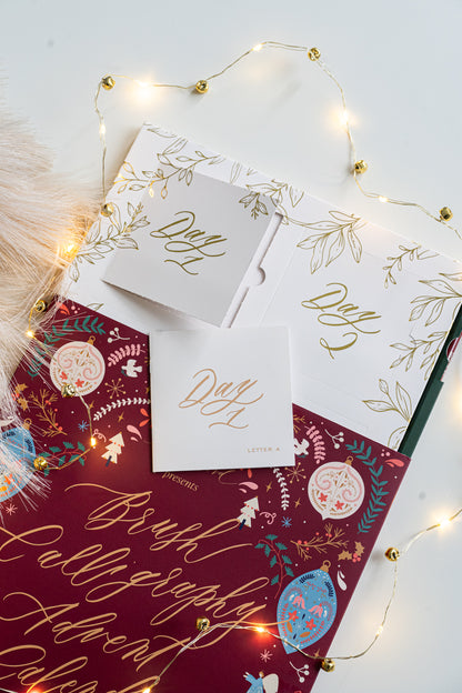Brush Calligraphy Advent Calendar opened to reveal the “Day 1” and “Day 2” gold foil doors, with the Day 1 booklet displayed on top of the festive maroon cover surrounded by twinkling fairy lights.
