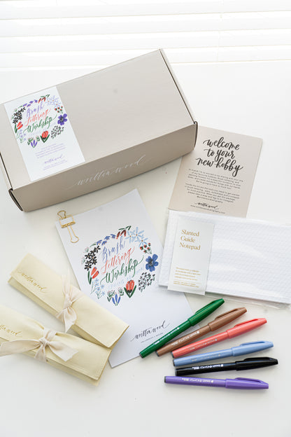 A brush lettering starter kit with a variety of items laid out, including a box with a floral design, brush pens, worksheets, and a notepad, all arranged on a white surface.
