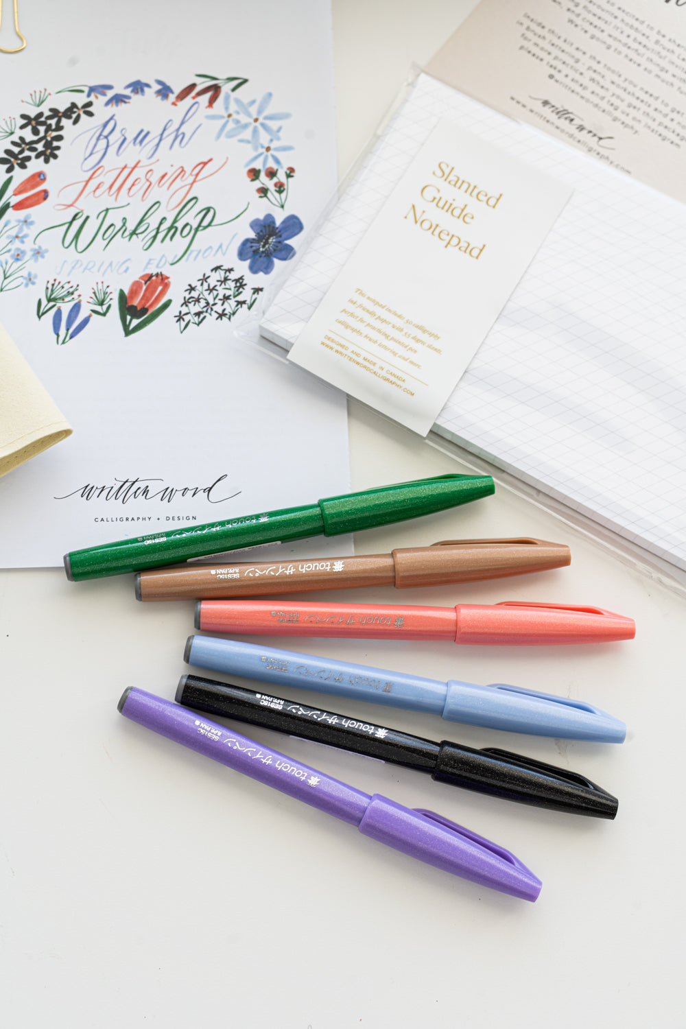 A brush lettering starter kit with a bundle of worksheets , six brush pens, and a notepad, all arranged on a white surface.