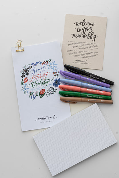 A brush lettering starter kit with a variety of items laid out, including a bundle of worksheets with a floral design title page, brush pens, and a notepad, all arranged on a white surface.