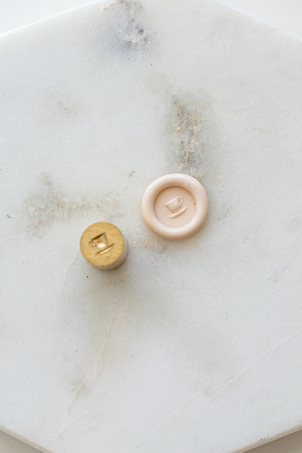 ​A mini brass wax seal stamp head with a 3D tea cup design beside a blush wax seal impression on a white marble surface