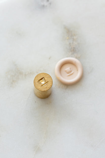 ​A mini brass wax seal stamp head with a 3D tea cup design beside a blush wax seal impression on a white marble surface