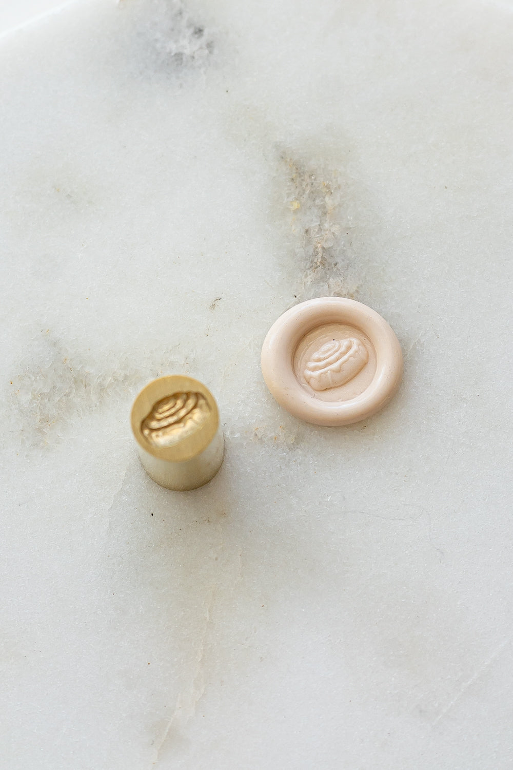 ​A mini brass wax seal stamp head with a 3D cinnamon bun design beside a blush wax seal impression on a white marble surface