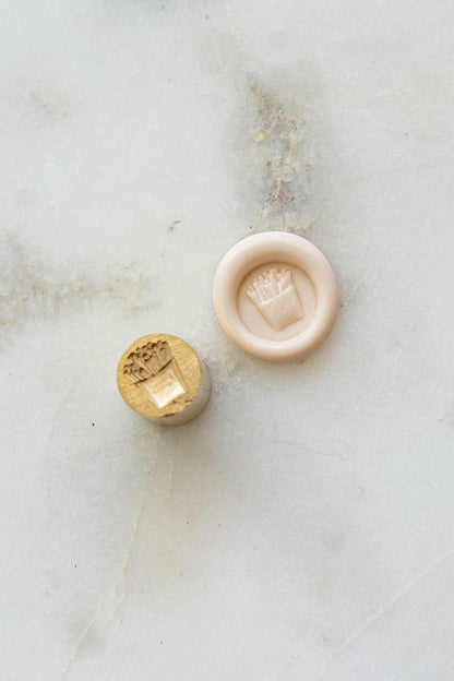 ​A mini brass wax seal stamp head with a 3D French fries design beside a blush wax seal impression on a white marble surface