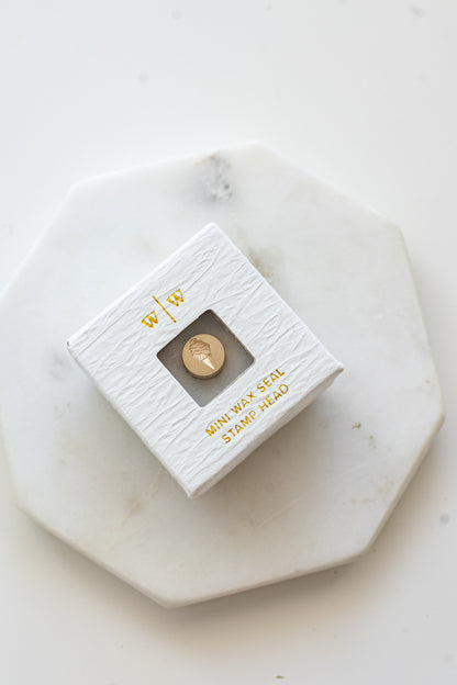 A mini brass wax seal stamp head with a 3D c design in a textured white ring-style box on a marble coaster