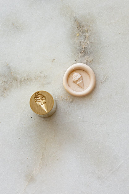 ​A mini brass wax seal stamp head with a 3D soft serve ice cream design beside a blush wax seal impression on a white marble surface