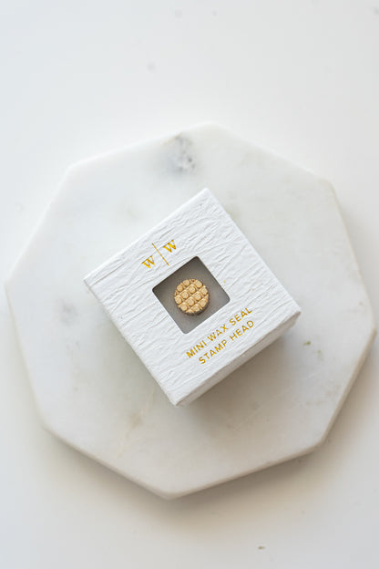 A mini brass wax seal stamp head with a 3D Belgian waffle design in a textured white ring-style box on a marble coaster