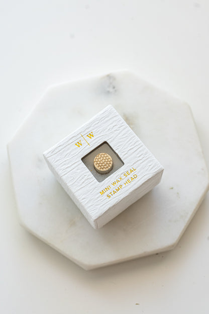 A mini brass wax seal stamp head with a 3D bubble waffle design in a textured white ring-style box on a marble coaster