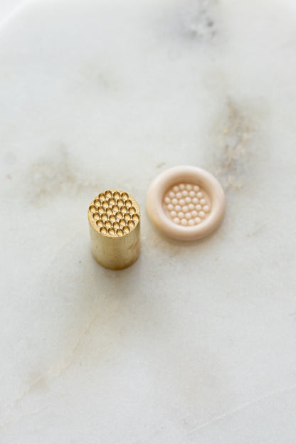 ​A mini brass wax seal stamp head with a 3D bubble waffle design beside a blush wax seal impression on a white marble surface