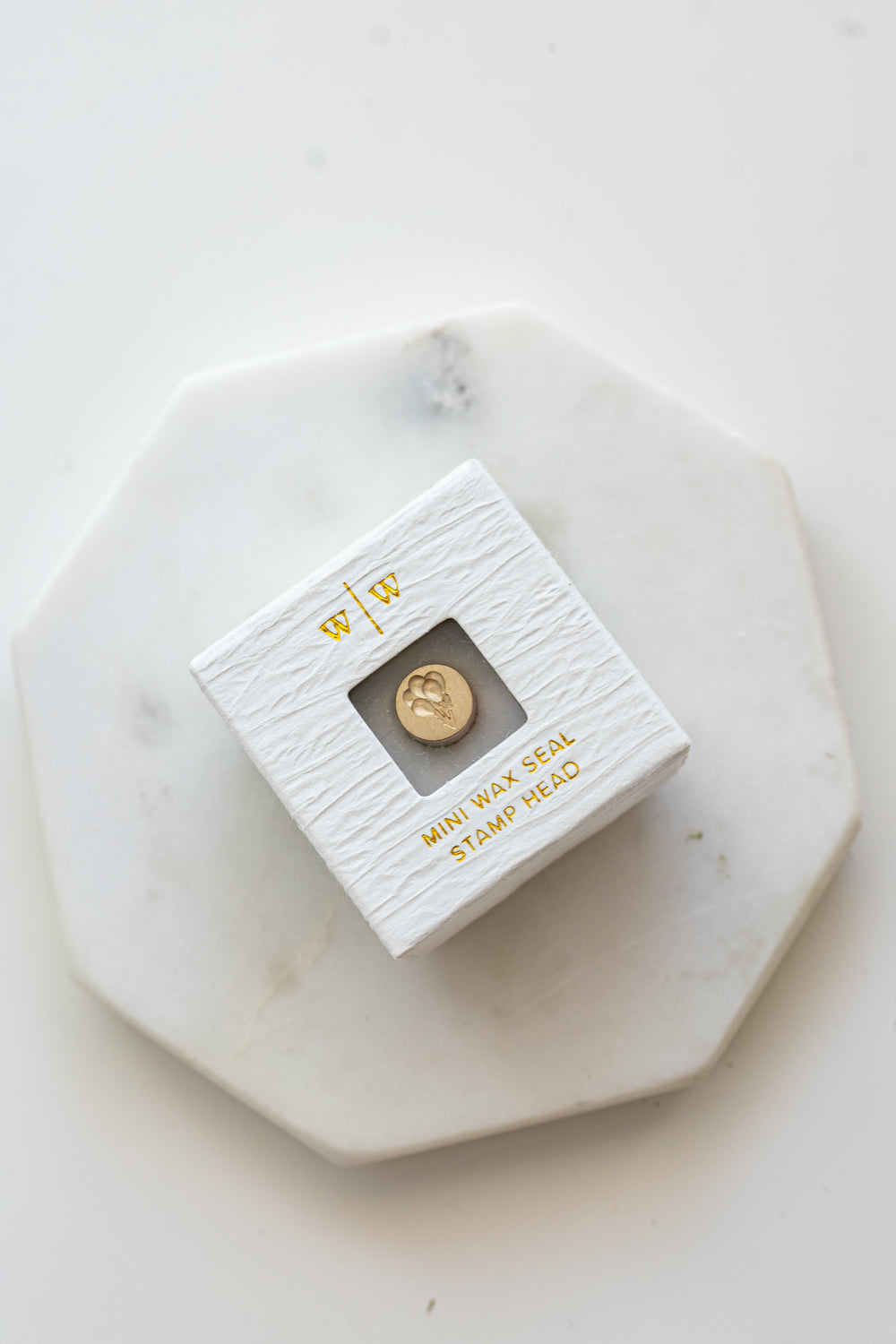 A mini brass wax seal stamp head with a 3D balloons design in a textured white ring-style box on a marble coaster