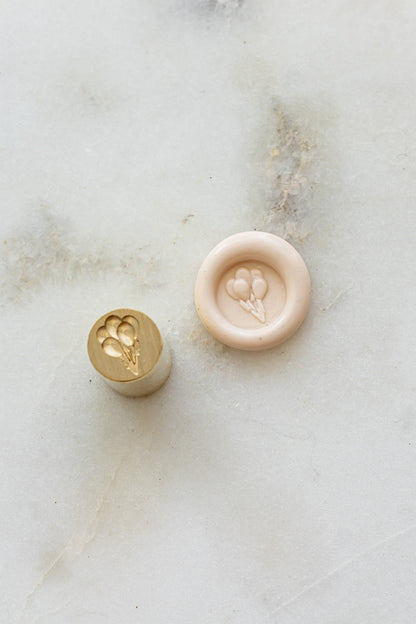 ​A mini brass wax seal stamp head with a 3D balloons design beside a blush wax seal impression on a white marble surface