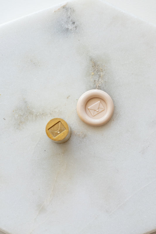​A mini brass wax seal stamp head with an envelope design beside a blush wax seal impression on a white marble surface
