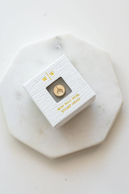 A mini brass wax seal stamp head with a 3D cupcake design in a textured white ring-style box on a marble coaster