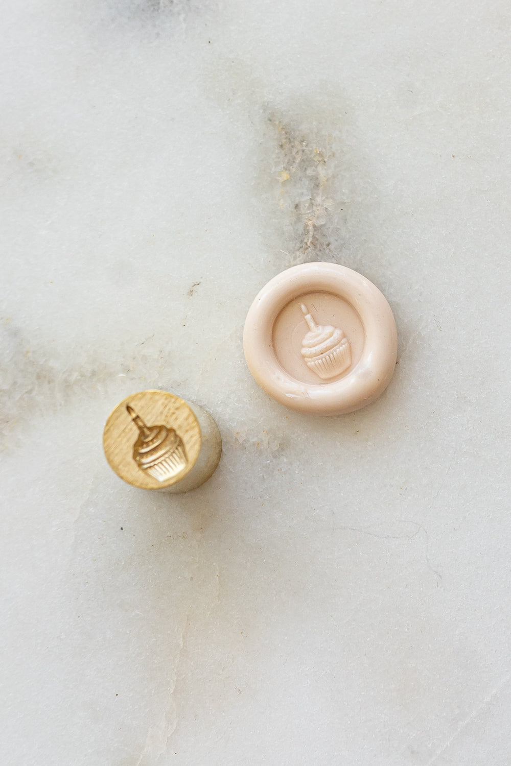 ​A mini brass wax seal stamp head with a 3D cupcake design beside a blush wax seal impression on a white marble surface