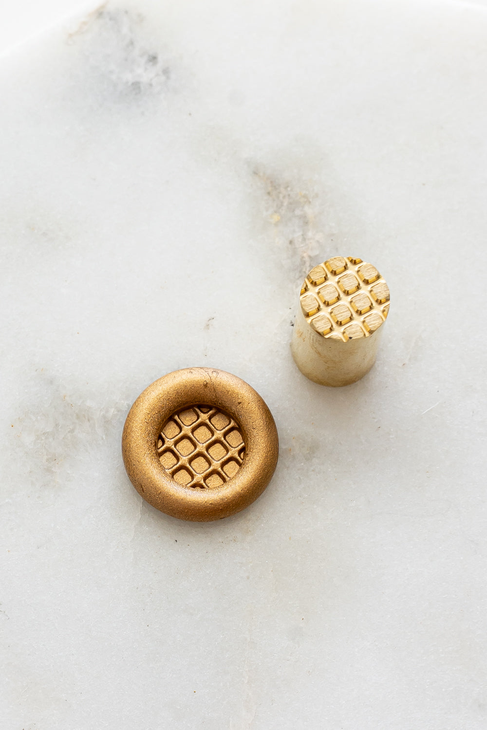 ​A mini brass wax seal stamp head with a 3D Belgian waffle design beside a gold wax seal impression on a white marble surface