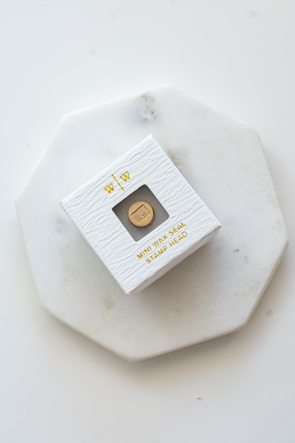 A mini brass wax seal stamp head with an envelope design in a textured white ring-style box on a marble coaster