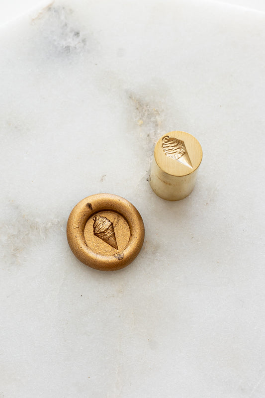 ​A mini brass wax seal stamp head with a 3D soft serve ice cream design beside a gold wax seal impression on a white marble surface