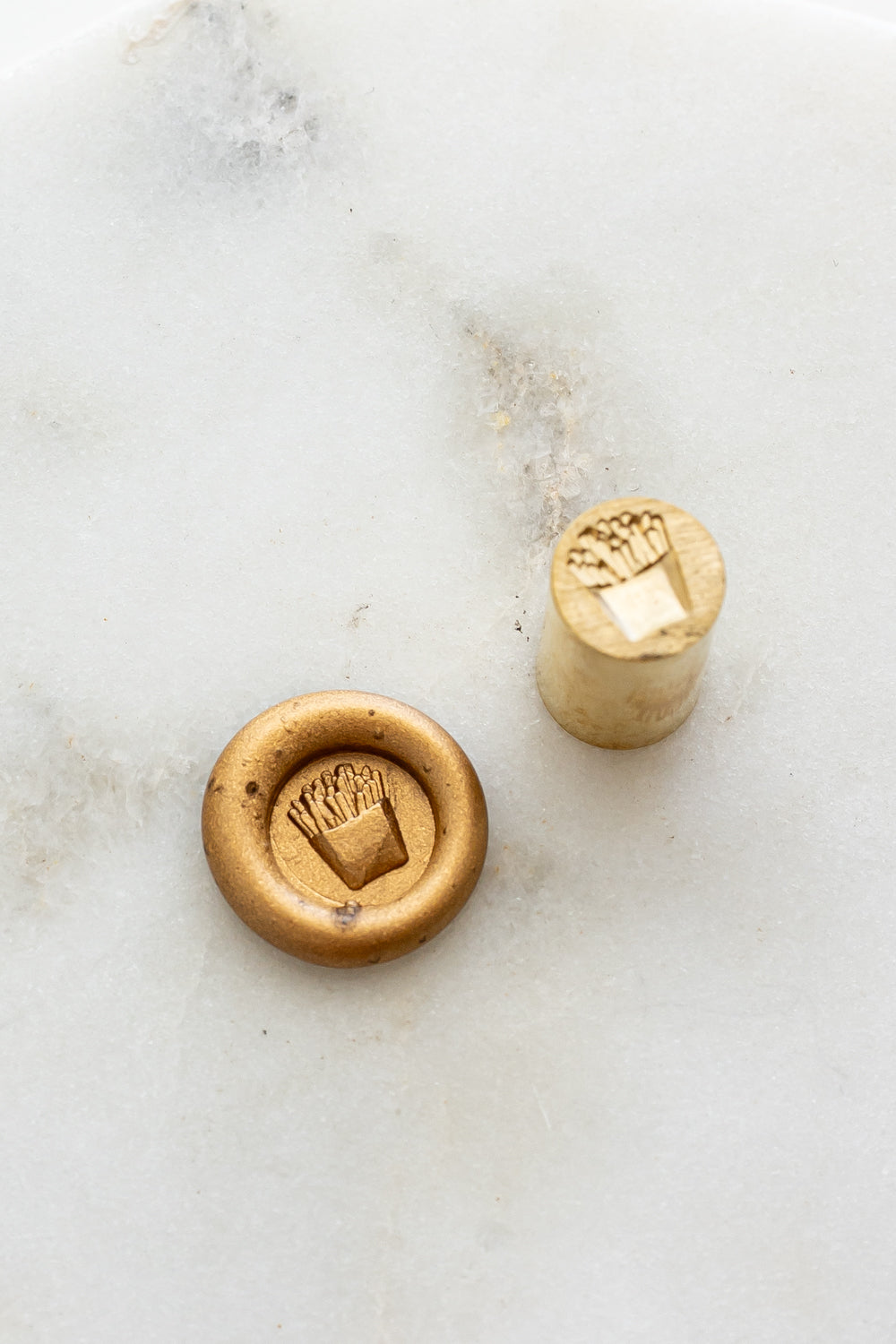 ​A mini brass wax seal stamp head with a 3D French fries design beside a gold wax seal impression on a white marble surface