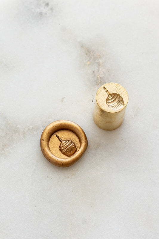 ​A mini brass wax seal stamp head with a 3D cupcake design beside a gold wax seal impression on a white marble surface