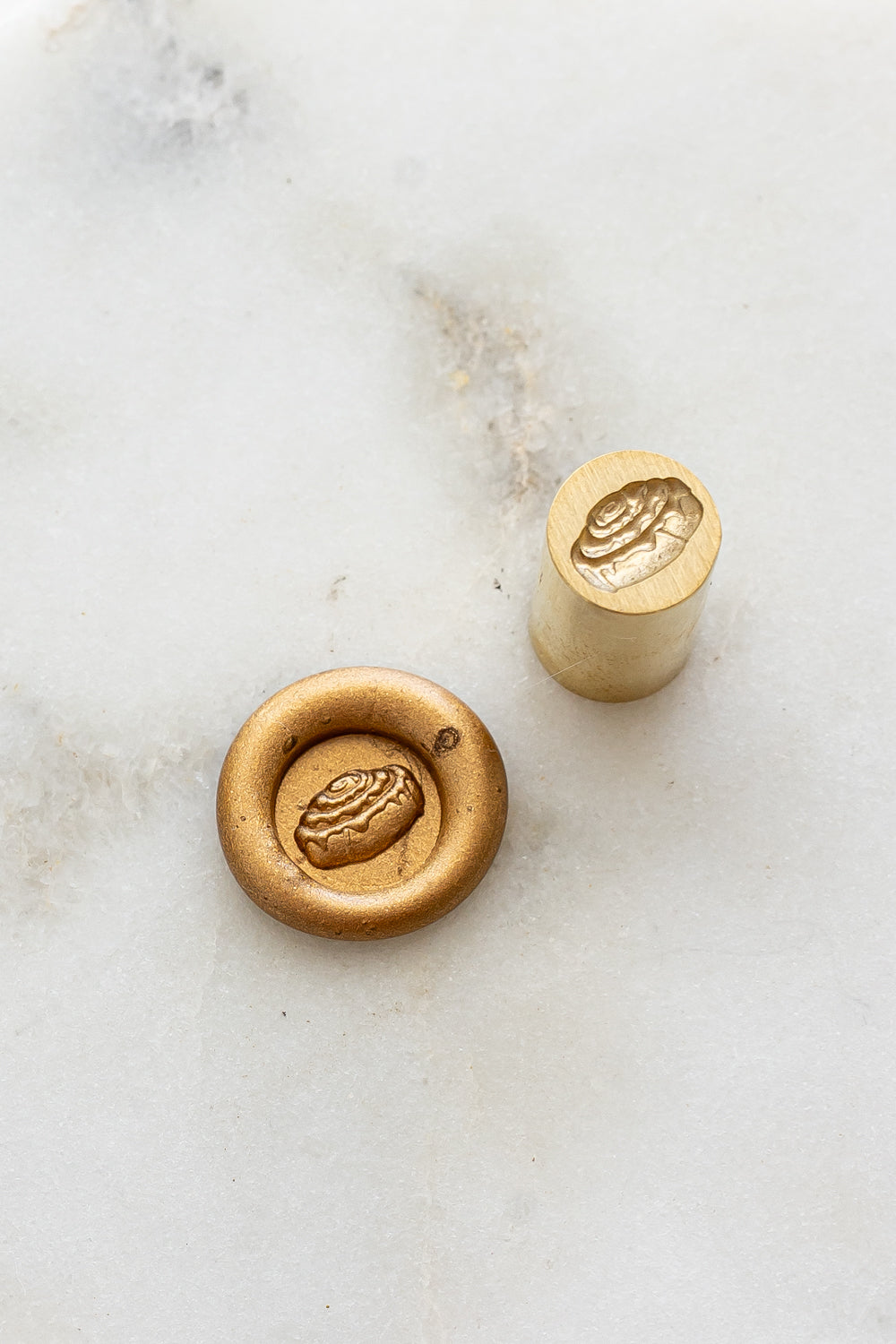 ​A mini brass wax seal stamp head with a 3D cinnamon bun design beside a gold wax seal impression on a white marble surface