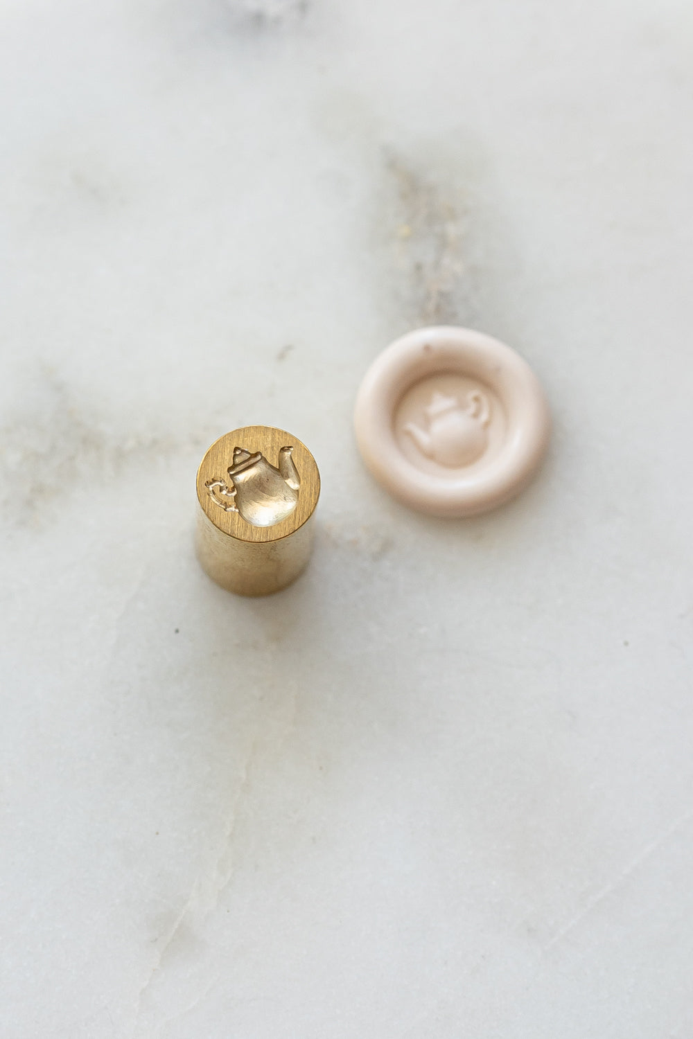 A mini brass wax seal stamp head with a 3D tea pot design beside a blush wax seal impression on a white marble surface