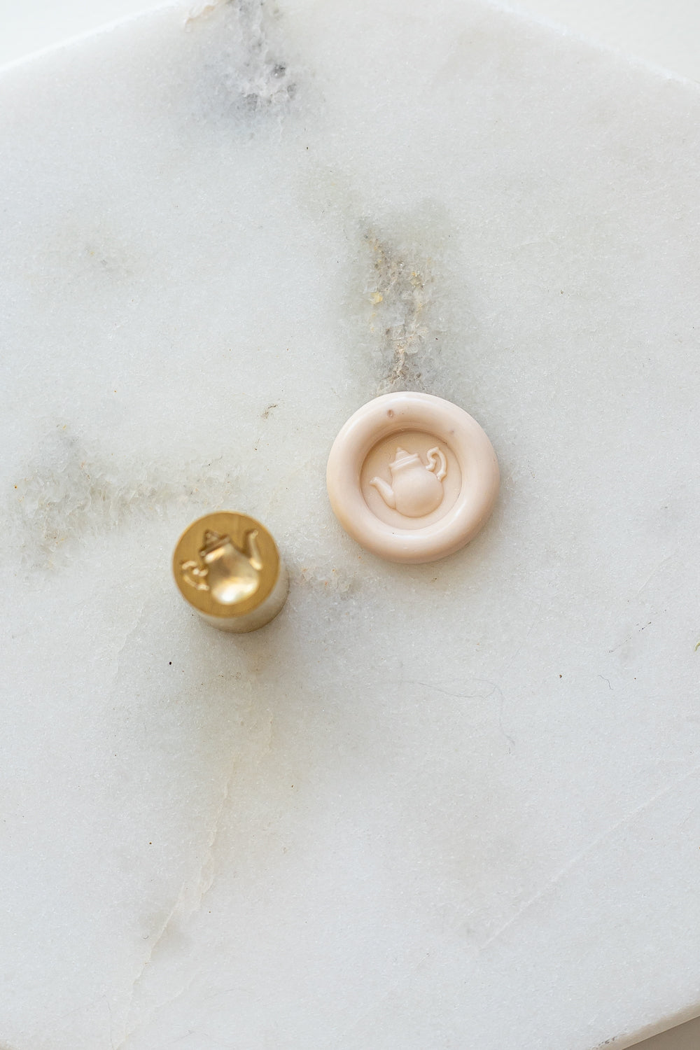A mini brass wax seal stamp head with a 3D tea pot design beside a blush wax seal impression on a white marble surface