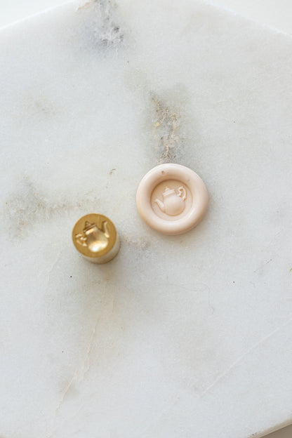A mini brass wax seal stamp head with a 3D tea pot design beside a blush wax seal impression on a white marble surface