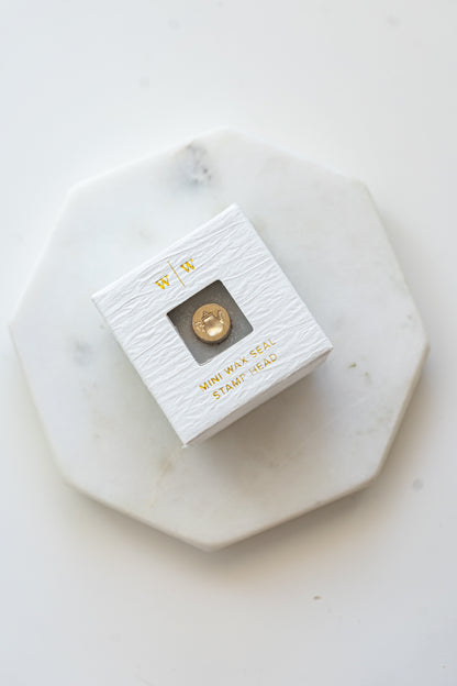 A mini brass wax seal stamp head with a 3D tea pot design in a textured white ring-style box on a marble coaster